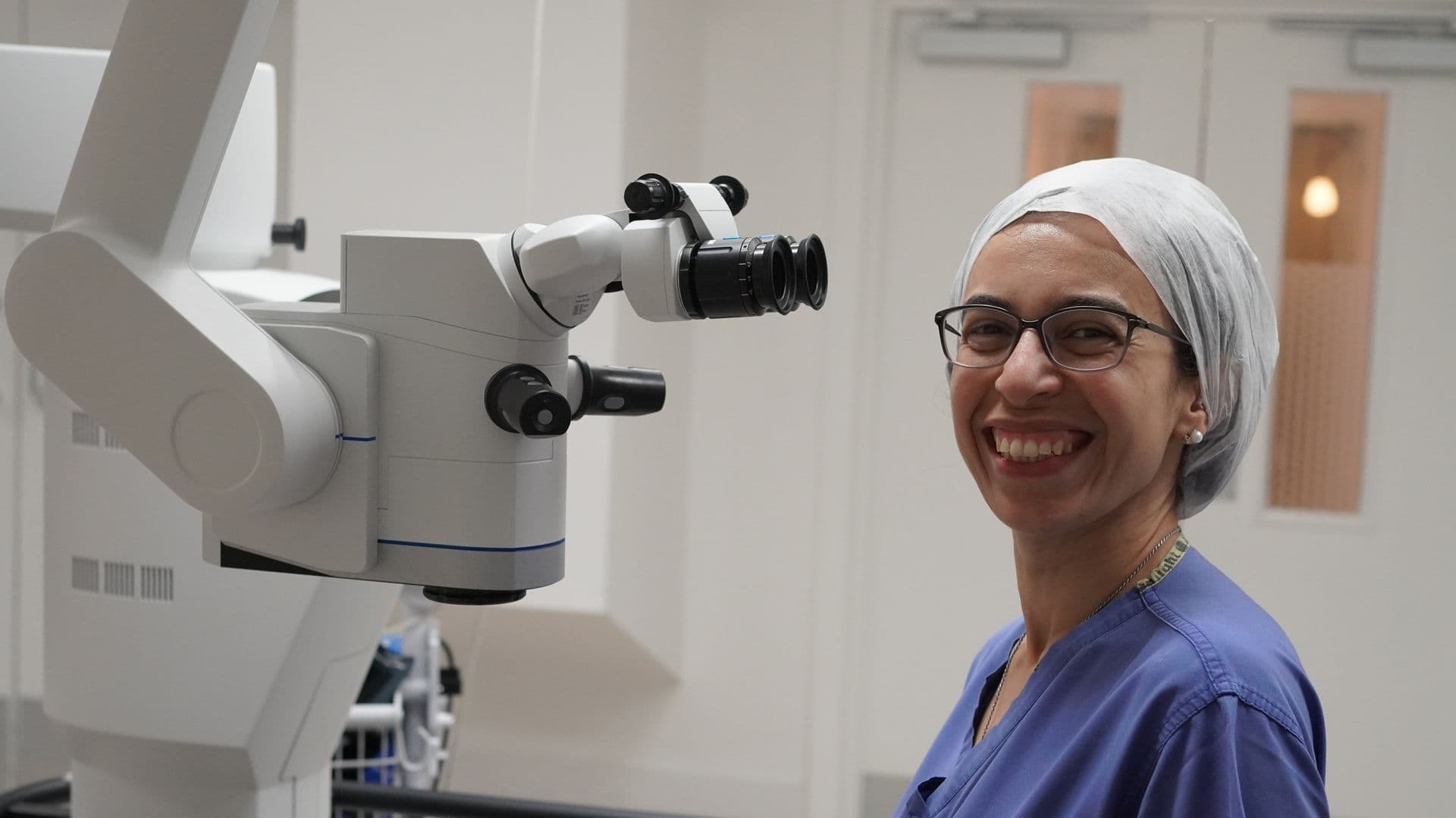 Miss Nardine Menassa, Consultant Ophthalmic Surgeon, smiling in surgical scrubs and a cap beside an operating microscope in the theatre at Spire Liverpool Hospital.
