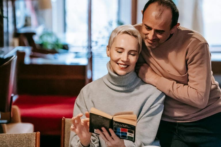 A woman reads a book while a man embraces her from behind in a cozy indoor setting.